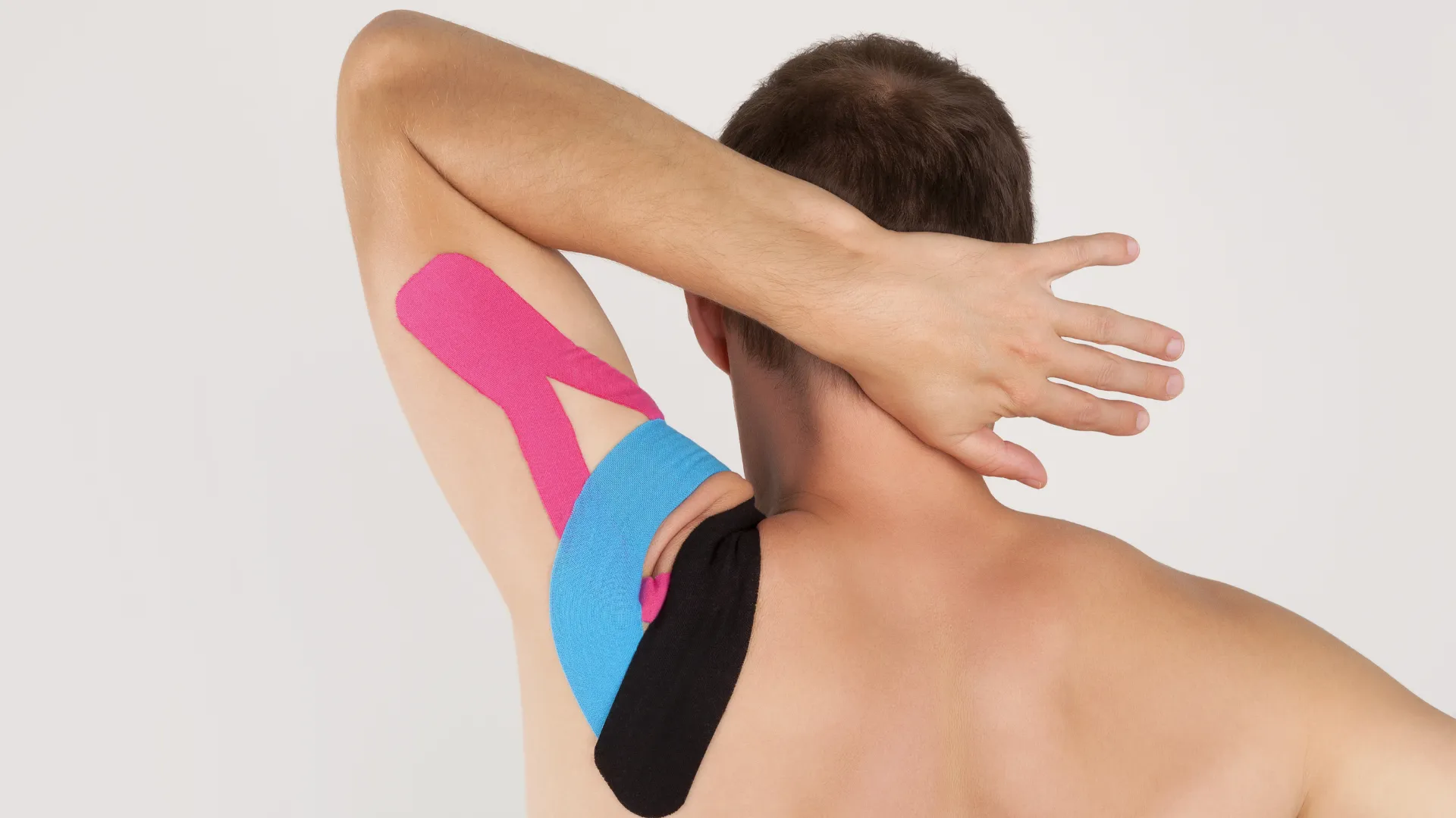 Hombre de espaldas levantando el brazo, con cintas de kinesiología de colores rosa, azul y negro aplicadas en el hombro y la axila sobre la piel desnuda.